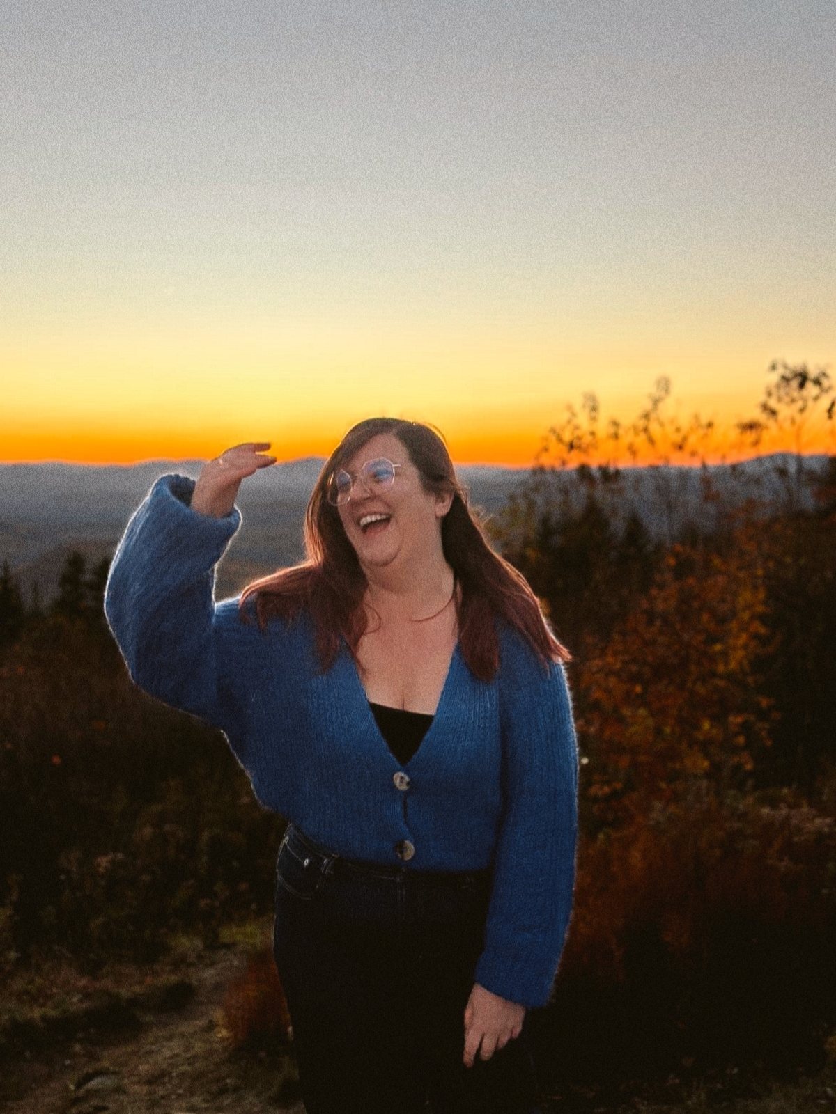 Photographie de Charline Couturier devant un coucher de soleil, portant un cardigan bleu tricoté main et levant la main pour remettre en place ses cheveux, un grand sourire aux lèvres.