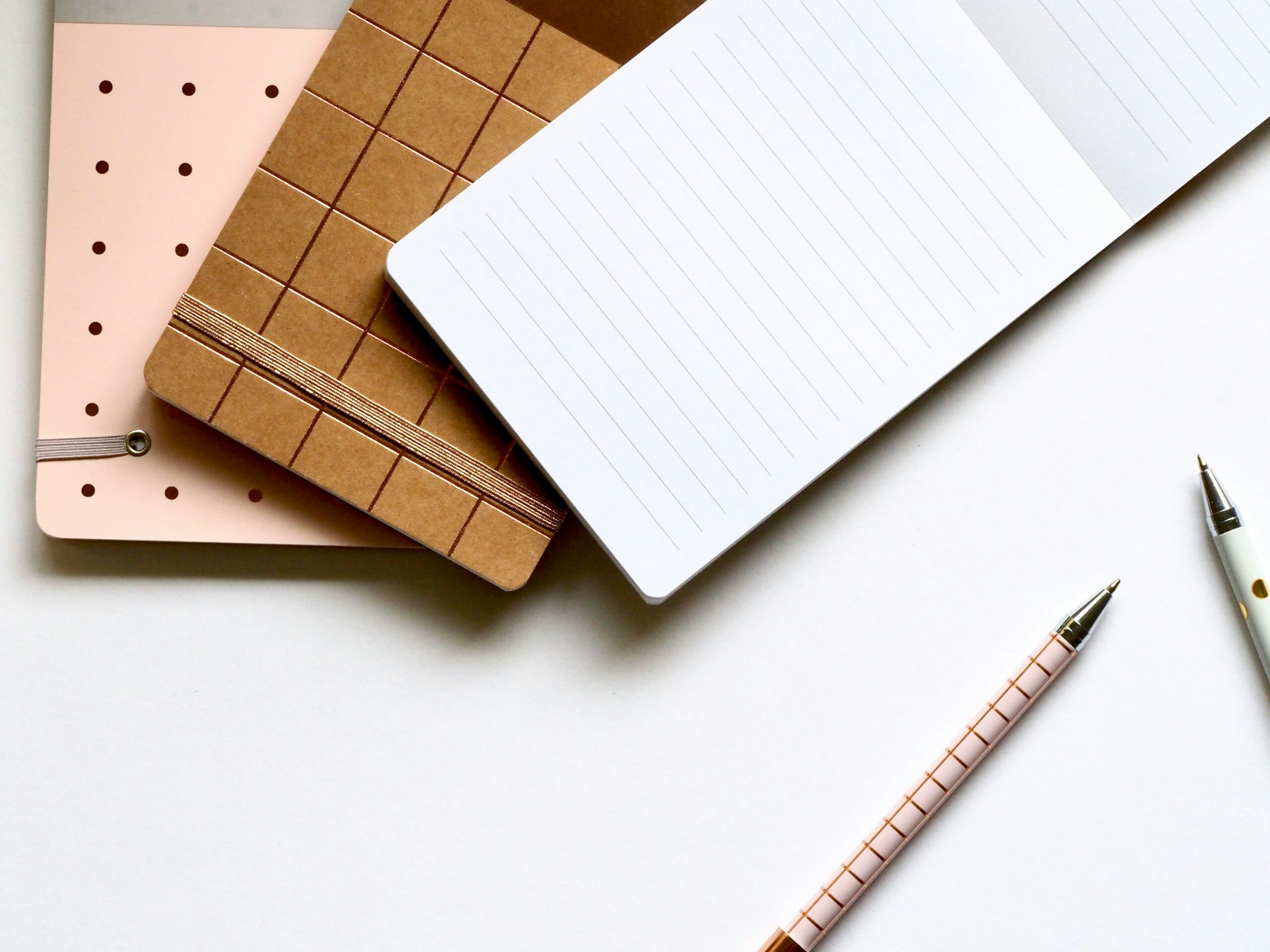 Trois carnets superposés sur une table blanche, accompagnés de deux stylos.